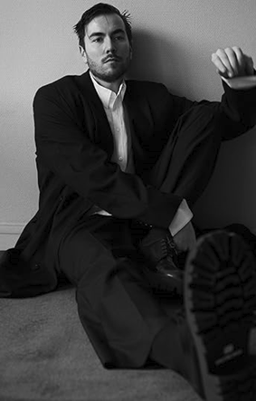 Black and white photo of author, Tomasz Jedrowski, sitting against a wall with one leg propped up.